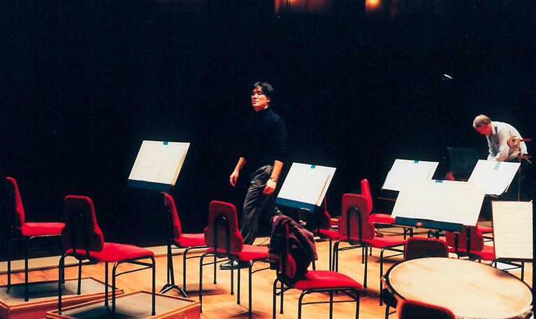 The conductor walking off the stage after a rehearsal, empty seats around him on the podium. Photograph.