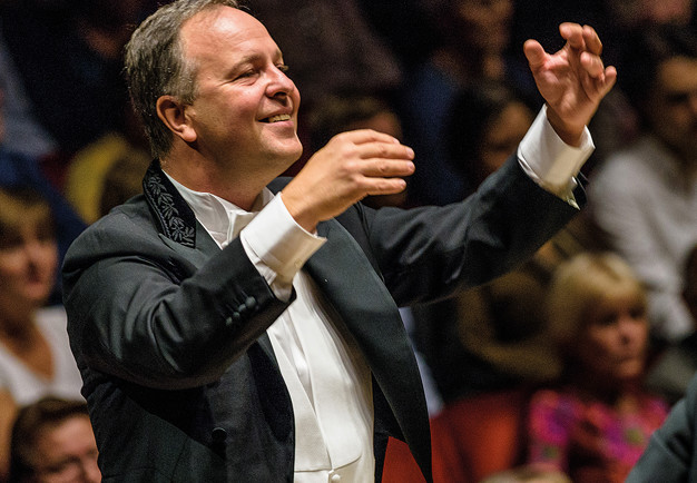 Smiling man conducting, audience can be seen behind him. Photography