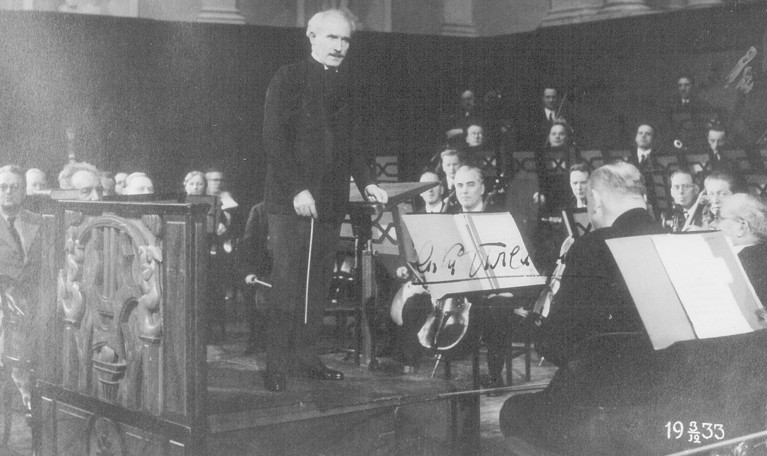 The conductor talking to the orchestra during rehearsal. Black and white photograph.